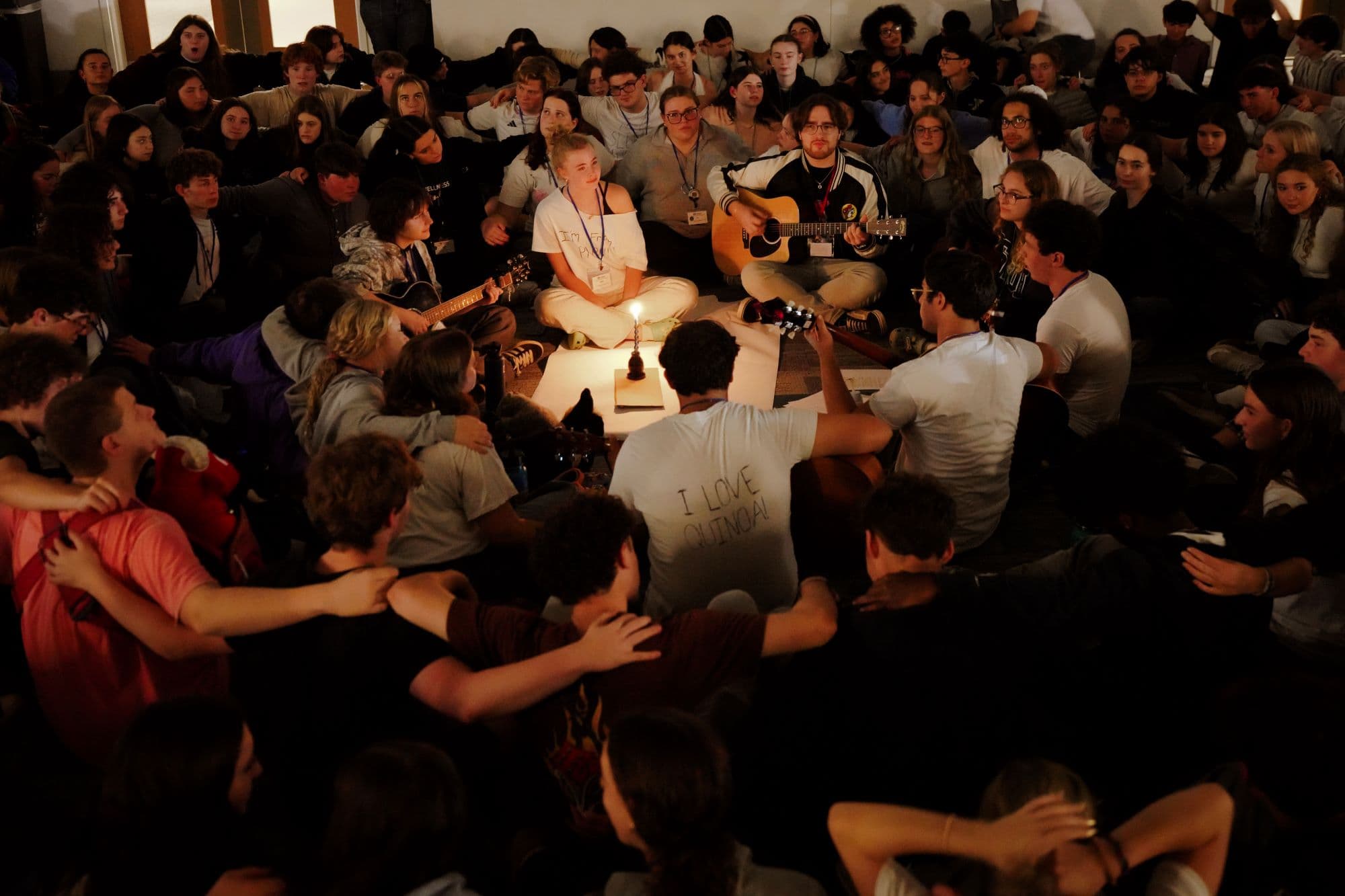 A group of teens gather during havdallah with their arms around one another. In the middle of the circle is a lit candle, shining light on hundreds of faces.