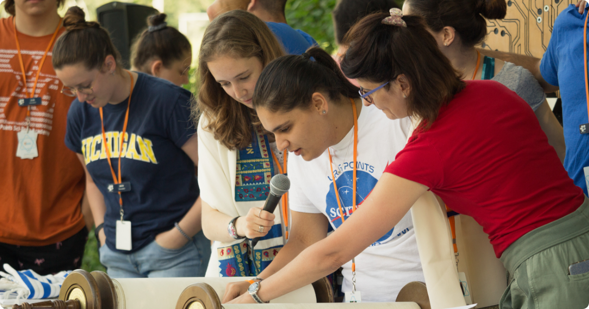 A camper reads torah while wearing a URJ 6 Points Sci-Tech shirt, surrounded by support around them.