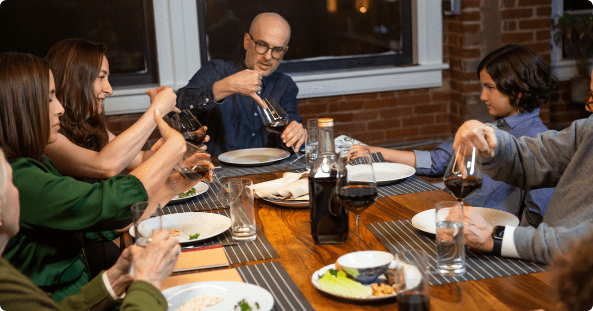 A family sits at the table holding a seder. They dip their pinkies in the wine glasses as part of the ritual.